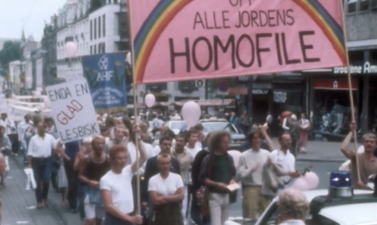 Folketog fra 70- eller 80-tallet, med fokus på en stor, rosa parole som viser en regnbue og ordene "Opp alle jordens homofile". En mindre parole med teksten "Enda en glad lesbisk" er også synlig i bakgrunnen.