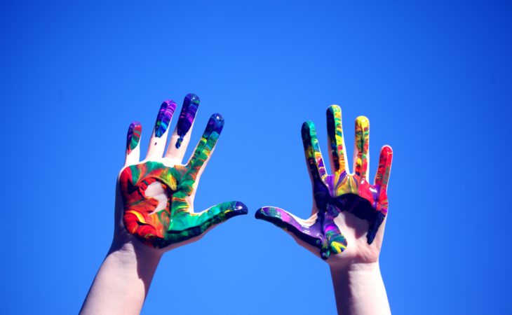 Bildet viser hendene til en person med lys hud som rekker opp mot en blå himmel. Hendene er helt dekket til med maling i alle regnbuens farger, som om de driver med fingermaling.